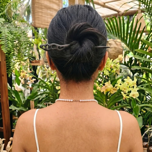 Épingle à Cheveux en Bois de Santal sculptée avec des motifs ajourés, maintenant le chignon d'une femme de dos dans un environnement tropical luxuriant.