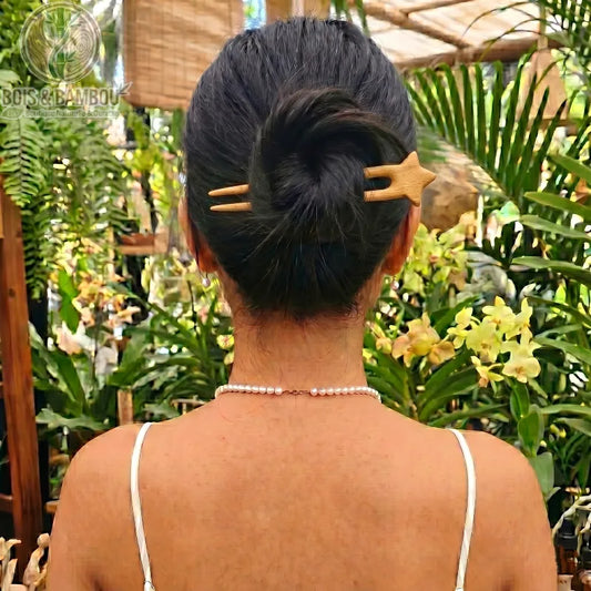 Épingle à Cheveux en U en Bois de Pêcher fixant le chignon d'une femme de dos, ornée d'un collier de perles dans un décor de serre tropicale.