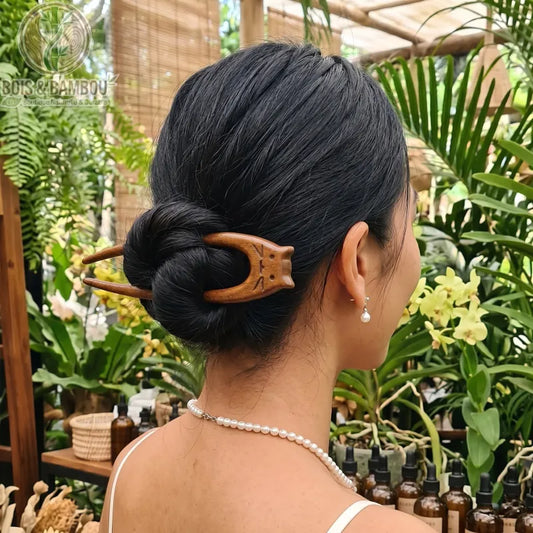 Épingle à cheveux chat en bois naturel sculptée en forme de tête de félin, fixant le chignon d'une femme de dos dans un décor de jardin tropical.