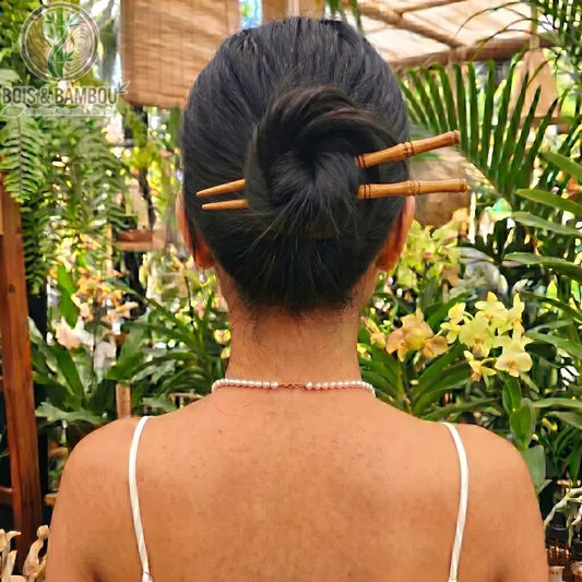 Épingles à Cheveux en Bois de Bambou en forme de tiges, maintenant le chignon d'une femme de dos avec un collier de perles dans un décor tropical.