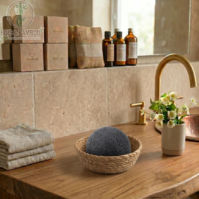 Éponge Konjac au charbon de bambou biologique. Une éponge noire demi-sphérique dans un panier tressé sur un meuble en bois, entourée d'un décor de salle de bain zen et naturel.