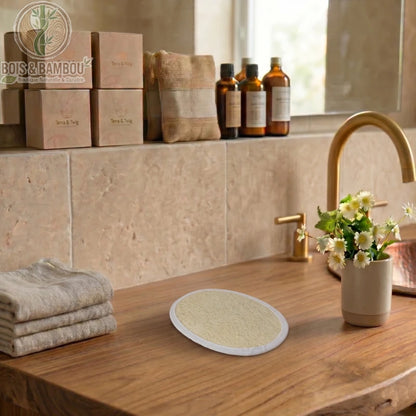 Éponge de luffa naturel de forme ovale et plate, posée sur un meuble de salle de bain en bois. Décor rustique avec serviettes, fleurs et flacons de soin pour une ambiance spa zen.