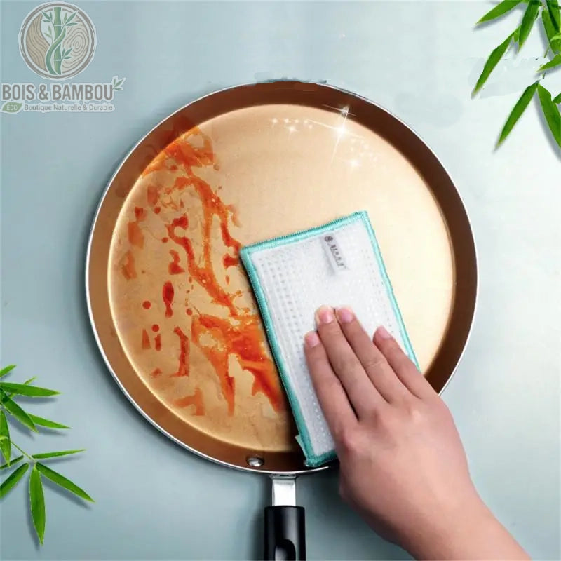 Éponge vaisselle en fibre de bambou. Une main utilisant une éponge rectangulaire blanche à bordure verte pour nettoyer les taches de graisse d'une poêle ronde dorée. Solution durable.