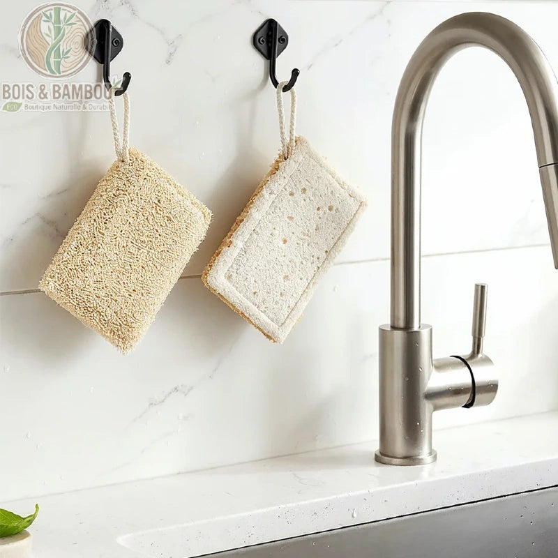 Éponges de Cuisine Eko. Deux éponges à récurer naturelles rectangulaires, l'une en luffa texturé et l'autre en cellulose plus douce, suspendues à des crochets noirs dans une cuisine moderne.