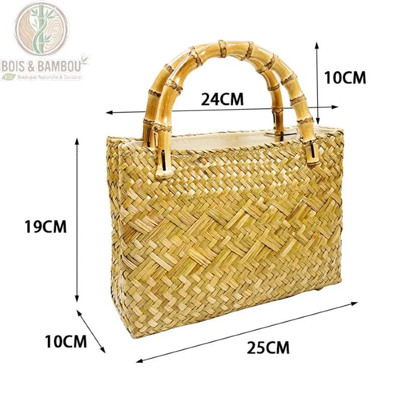 Fourre tout en paille tressee situation 3. Une femme en robe en jean tenant un sac à main rectangulaire en paille tressée dorée avec des anses en bambou, assise sur des marches en pierre.
