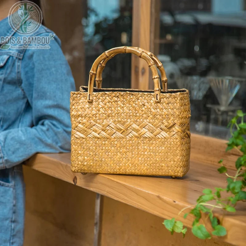 Fourre tout en paille tressee situation 2. Sac artisanal de forme rectangulaire en paille tressée, avec deux anses en bambou courbé, posé sur une étagère en bois dans une boutique à l'ambiance naturelle.