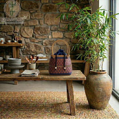 Grand fourre tout Nomade en bambou 4. Panier artisanal en bambou bicolore avec anse haute et pochon bleu marine, exposé sur un banc rustique dans un décor naturel avec mur en pierre et plante verte.
