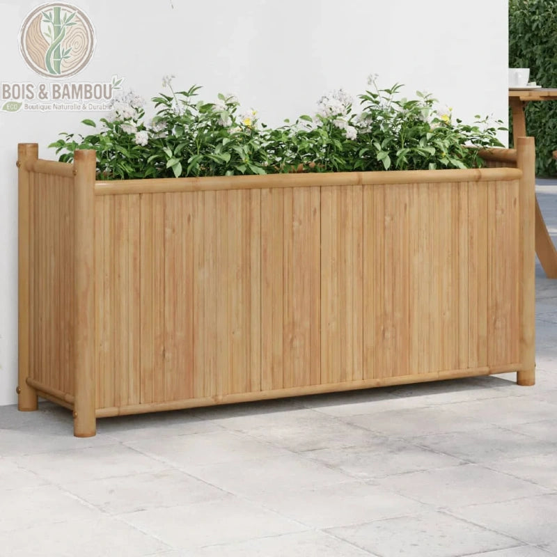 Jardinière en Bambou 100 cm. Un bac à fleurs rectangulaire robuste en bambou clair, orné de plantes fleuries blanches, posé sur une terrasse dallée devant un mur blanc épuré.