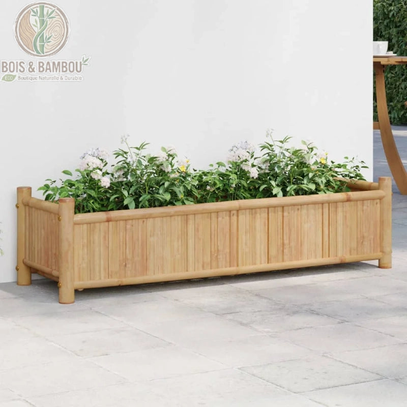 Jardinière en Bambou Naturel. Une grande jardinière rectangulaire en bambou clair remplie de plantes aux fleurs blanches, posée sur une terrasse en dalles devant un mur blanc.