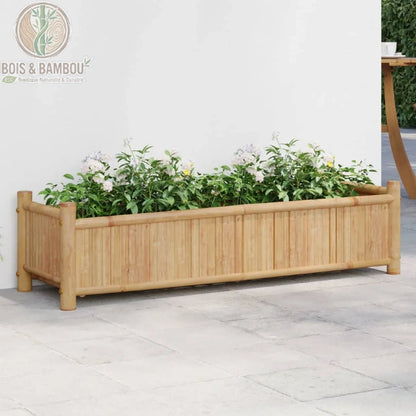 Jardinière en Bambou Naturel. Une grande jardinière rectangulaire en bambou clair remplie de plantes aux fleurs blanches, posée sur une terrasse en dalles devant un mur blanc.