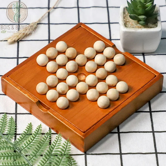 Jeu de Solitaire en bois. Plateau carré en bois verni avec des billes en bois clair disposées en forme de croix, posé sur une nappe quadrillée à côté d'une petite plante.