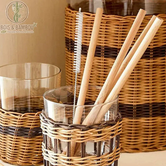 Kit de 12 pailles en bambou bio. Trois pailles en bois naturel et un goupillon de nettoyage dans un verre habillé d'osier tressé, devant une carafe assortie. Style bohème et durable.