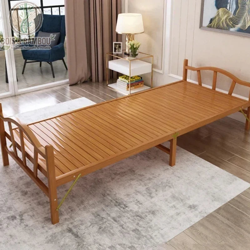Lit Pliant en Bambou Massif 100x192. Lit simple pliant en bambou massif verni avec tête et pied de lit cintrés, disposé dans une chambre claire sur un tapis gris, près d'un guéridon de chevet design.