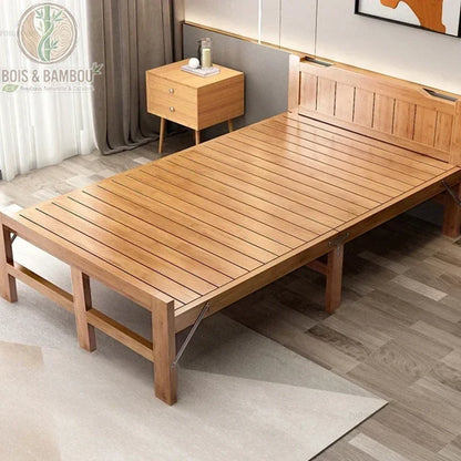Lit Pliant en Bambou Massif 100x207. Lit simple en bambou massif verni avec sommier à lattes et tête de lit, installé sur un tapis beige dans une chambre moderne à côté d'un chevet en bois.