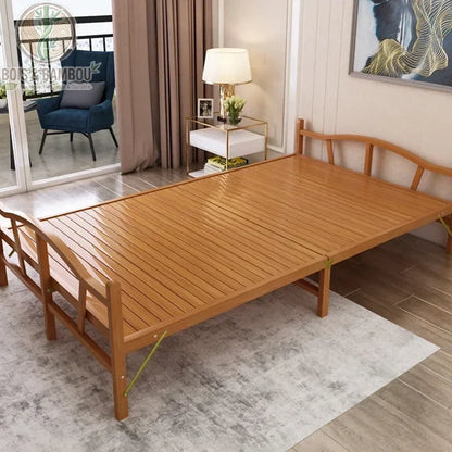 Lit Pliant en Bambou Massif 120x192. Lit d'appoint en bambou massif verni avec tête et pied de lit arqués, installé sur un tapis gris dans une chambre lumineuse avec vue sur un balcon.