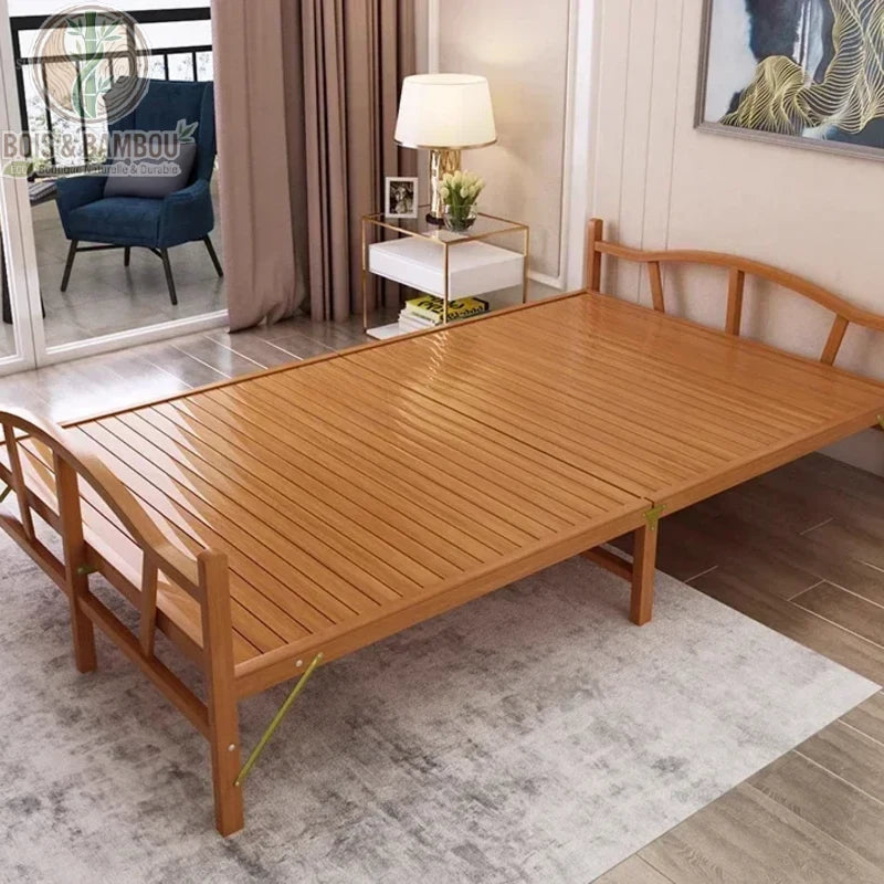 Lit Pliant en Bambou Massif 150x192 2. Large lit double pliant en bambou massif verni avec tête et pied de lit arqués, installé sur un tapis gris dans une chambre lumineuse avec vue sur un balcon.