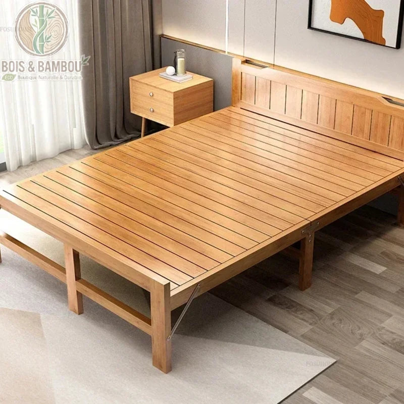 Lit Pliant en Bambou Massif 150x197. Large lit double pliant en bambou massif verni avec sommier à lattes, présenté dans une chambre moderne à côté d'une table de chevet assortie sur un tapis clair.