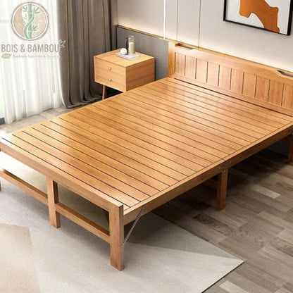 Lit Pliant en Bambou Massif 150x197. Large lit double pliant en bambou massif verni avec sommier à lattes, présenté dans une chambre moderne à côté d'une table de chevet assortie sur un tapis clair.