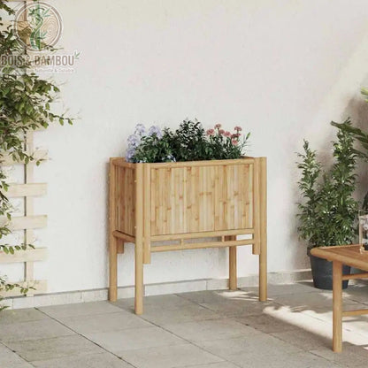 Lit Surélevé en Bambou. Un bac de plantation rectangulaire sur pieds en bambou clair, rempli de fleurs violettes et roses, installé sur une terrasse contre un mur blanc.