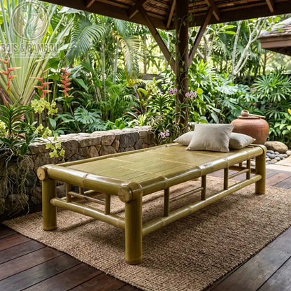 Lit en Bambou Tressé Carbonisé Classique. Un lit de repos robuste en bambou épais avec plateau tressé et coussins, installé sous un pavillon ouvert entouré d'un jardin tropical.