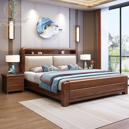 Lit en Noyer de Soie Dorée. Un lit luxueux en bois massif foncé avec une tête de lit rembourrée beige intégrant une niche de rangement, entouré de deux tables de chevet assorties.