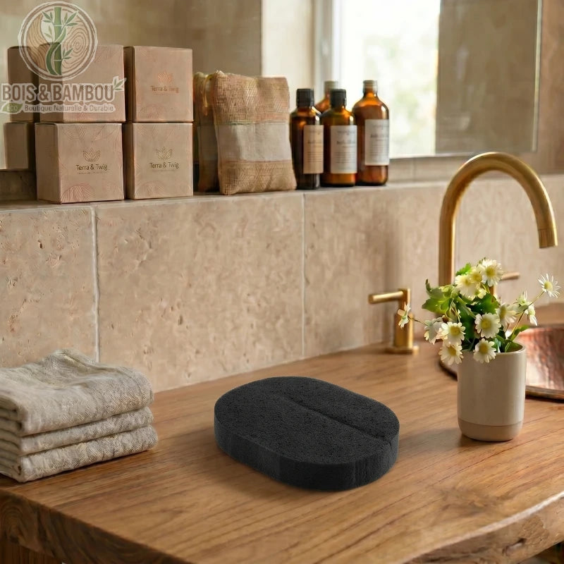 Lot de 2 éponges faciales au charbon de bambou. Une éponge noire ovale et une luffa fibreuse pliée sur un plan de travail en bois. Décor de salle de bain zen avec fleurs et flacons.