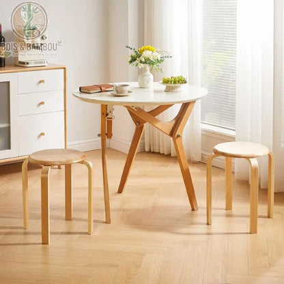 Lot de 4 Tabourets en Bois Massif situation. Deux tabourets en bois clair entourent une table ronde blanche avec des décorations, dans une pièce lumineuse avec parquet et meuble blanc.
