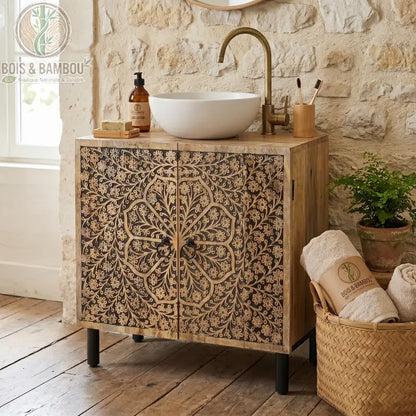 Meuble Sous Évier en Manguier : meuble élégant en bois massif avec façades sculptées de motifs floraux artisanaux dans une salle de bain avec un mur en pierres blanches. Comprend une découpe pour évier et des pieds en métal noir.