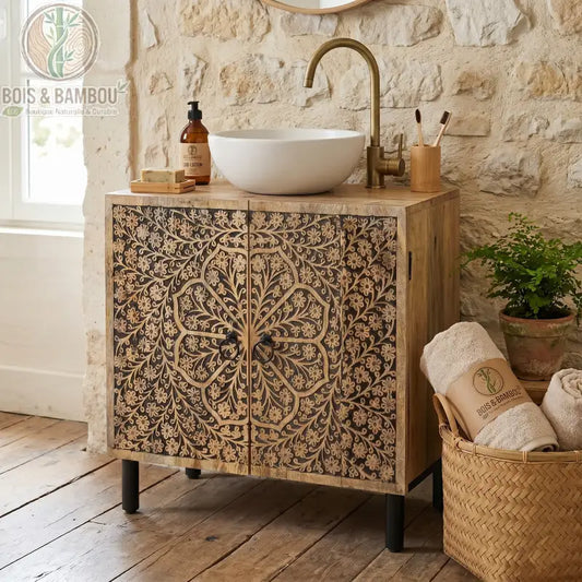 Meuble Sous Évier en Manguier : meuble élégant en bois massif avec façades sculptées de motifs floraux artisanaux dans une salle de bain avec un mur en pierres blanches. Comprend une découpe pour évier et des pieds en métal noir.