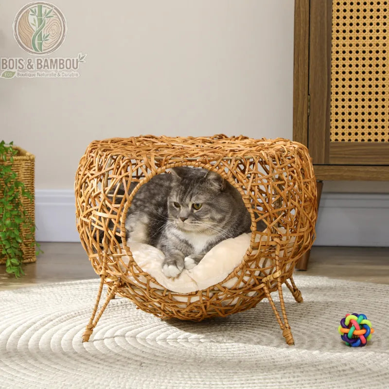 Niche pour Chat en Rotin. Un chat tigré gris repose confortablement sur un coussin moelleux à l'intérieur d'une niche sphérique sur pieds en rotin tressé, posée sur un tapis rond.