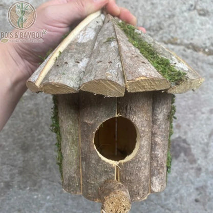 Nichoir de jardin rustique en bois. Un abri artisanal en écorce naturelle avec un toit conique recouvert de mousse et un petit perchoir, tenu à la main sur un fond en pierre.