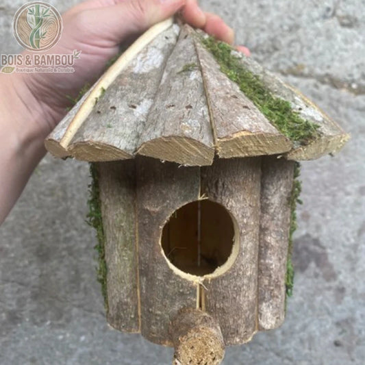 Nichoir de jardin rustique en bois. Un abri artisanal en écorce naturelle avec un toit conique recouvert de mousse et un petit perchoir, tenu à la main sur un fond en pierre.