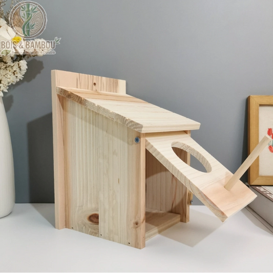 Nichoir extérieur en bois massif. Un nichoir rectangulaire en bois clair avec un toit en pente et une façade basculante percée d'un trou d'entrée circulaire munie d'un perchoir.