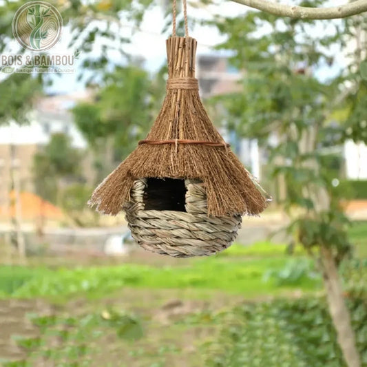Nid d'oiseau en paille tissée. Un petit abri artisanal suspendu, composé d'une base en paille tressée et d'un toit pointu en chaume, accroché à une branche dans un jardin.
