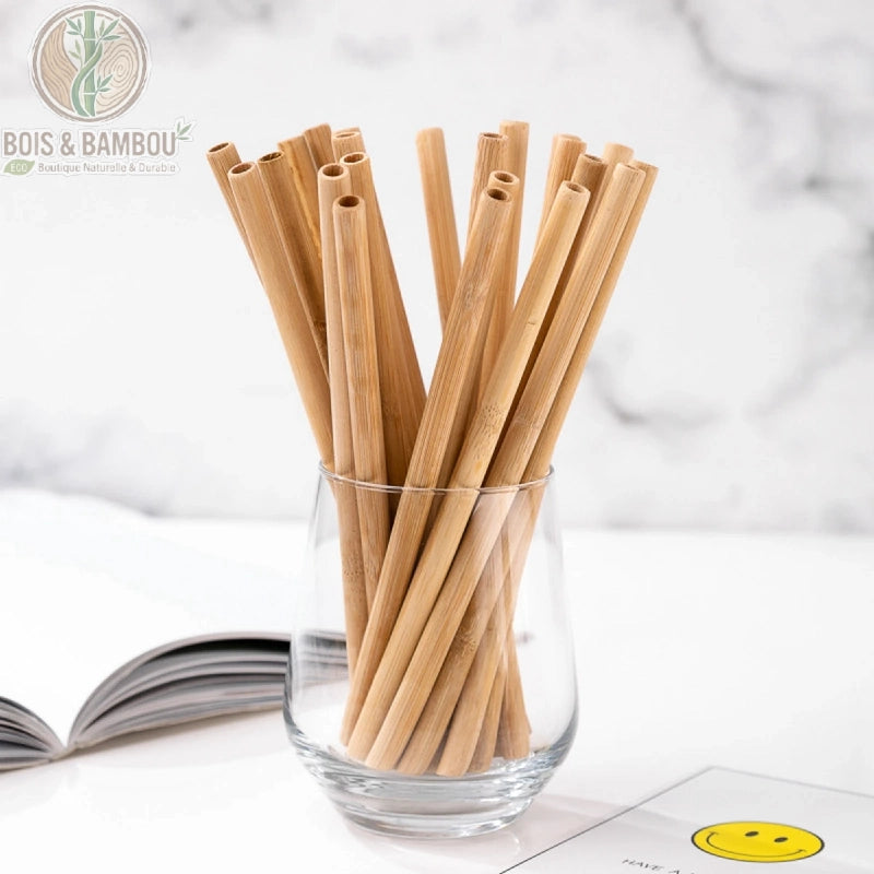 Pailles en Bambou Naturel. Un bouquet de pailles réutilisables en bambou clair disposées dans un verre transparent sur une table blanche. Alternative écologique et durable.