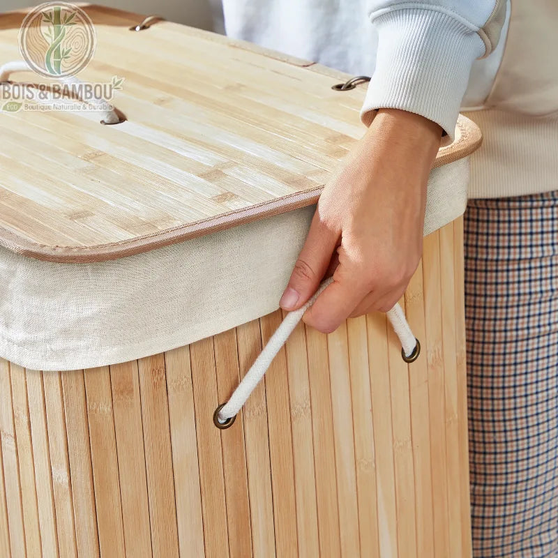 Panier a Linge Bambou Naturel avec Couvercle 72L situation 1. Gros plan sur un panier en bambou clair avec son couvercle assorti et sa poignée en corde blanche tenue par une main.
