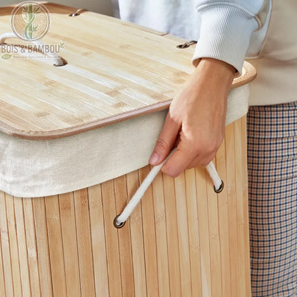 Panier a Linge Bambou Naturel avec Couvercle 72L situation 1. Gros plan sur un panier en bambou clair avec son couvercle assorti et sa poignée en corde blanche tenue par une main.