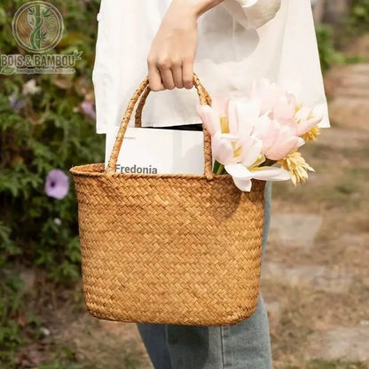 Panier en bambou tresse situation 1. Une personne porte à la main un sac cabas en bambou tressé naturel contenant des fleurs roses et un magazine, lors d'une promenade printanière en extérieur.