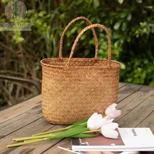 Panier en bambou tresse situation 2. Un sac cabas souple en bambou tressé naturel posé sur une table en bois extérieure, accompagné de tulipes blanches et d'un magazine sur fond de verdure.