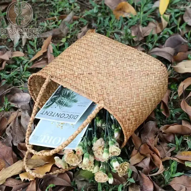 Panier en bambou tresse situation 3. Un sac cabas en bambou tressé renversé sur un lit de feuilles mortes, laissant échapper des fleurs fraîches et des magazines, pour une mise en scène champêtre.
