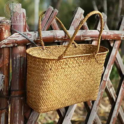 Panier en bambou tresse situation 4. Un cabas artisanal en bambou tressé suspendu à une clôture rustique en bois, illustrant son aspect pratique et naturel pour une utilisation en extérieur.
