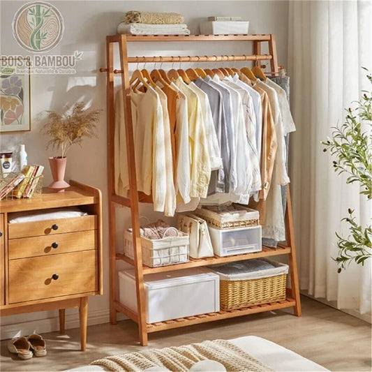 Penderie Bambou Naturel avec etageres et Crochets. Un portant en bois garni de vêtements suspendus et de paniers de rangement sur ses étagères inférieures, dans une chambre lumineuse et ordonnée.