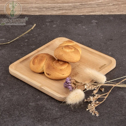 Petit Plateau Carré en Bambou. Un plateau carré en bambou clair avec des bords arrondis, présentant des petits pains dorés et des fleurs séchées sur une surface sombre texturée.