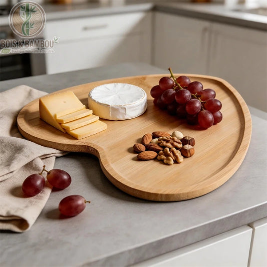 Plateau Cœur en Bambou. Un plateau en bambou clair en forme de cœur garni de fromages, de raisins rouges et de noix, posé sur un plan de travail gris dans une cuisine lumineuse.
