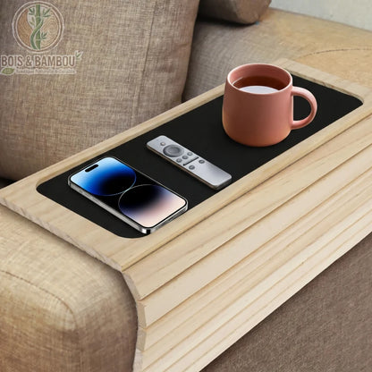 Plateau d accoudoir de canape en bambou 2. Un plateau flexible en bois clair sur un canapé, doté d'un revêtement noir antidérapant avec un smartphone, une télécommande et une tasse de thé rose.