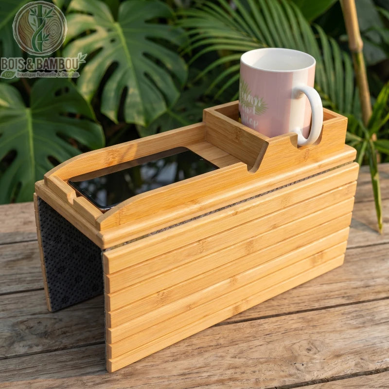 Plateau d accoudoir de canape en bambou 3. Un plateau flexible en bambou naturel posé sur du bois, doté d'un support pour smartphone et d'un compartiment avec une tasse rose, sur fond de verdure.