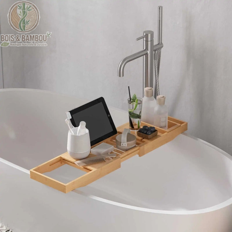 Pont de baignoire en bambou réglable. Un plateau extensible en bois clair posé sur une baignoire blanche, équipé d'un support pour tablette, d'un verre et d'accessoires de bien-être.