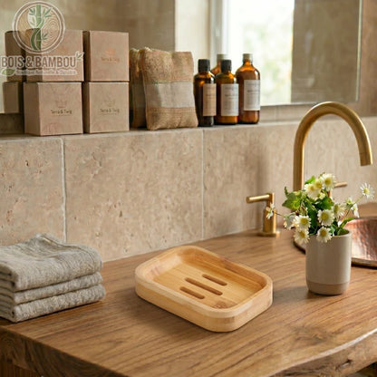 Porte-Savon en Bambou Naturel sur plan de travail en bois. Porte-savon rectangulaire fendu à côté de serviettes beiges et de fleurs blanches dans une salle de bain zen et rustique.