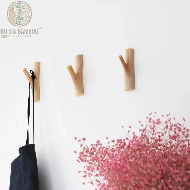 Porte manteau en bois massif mural situation. Trois patères en forme de branches naturelles fixées au mur, l'une portant un sac noir, avec un bouquet de fleurs roses séchées au premier plan.