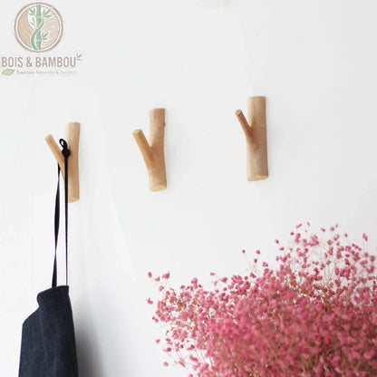 Porte manteau en bois massif mural situation. Trois patères en forme de branches naturelles fixées au mur, l'une portant un sac noir, avec un bouquet de fleurs roses séchées au premier plan.